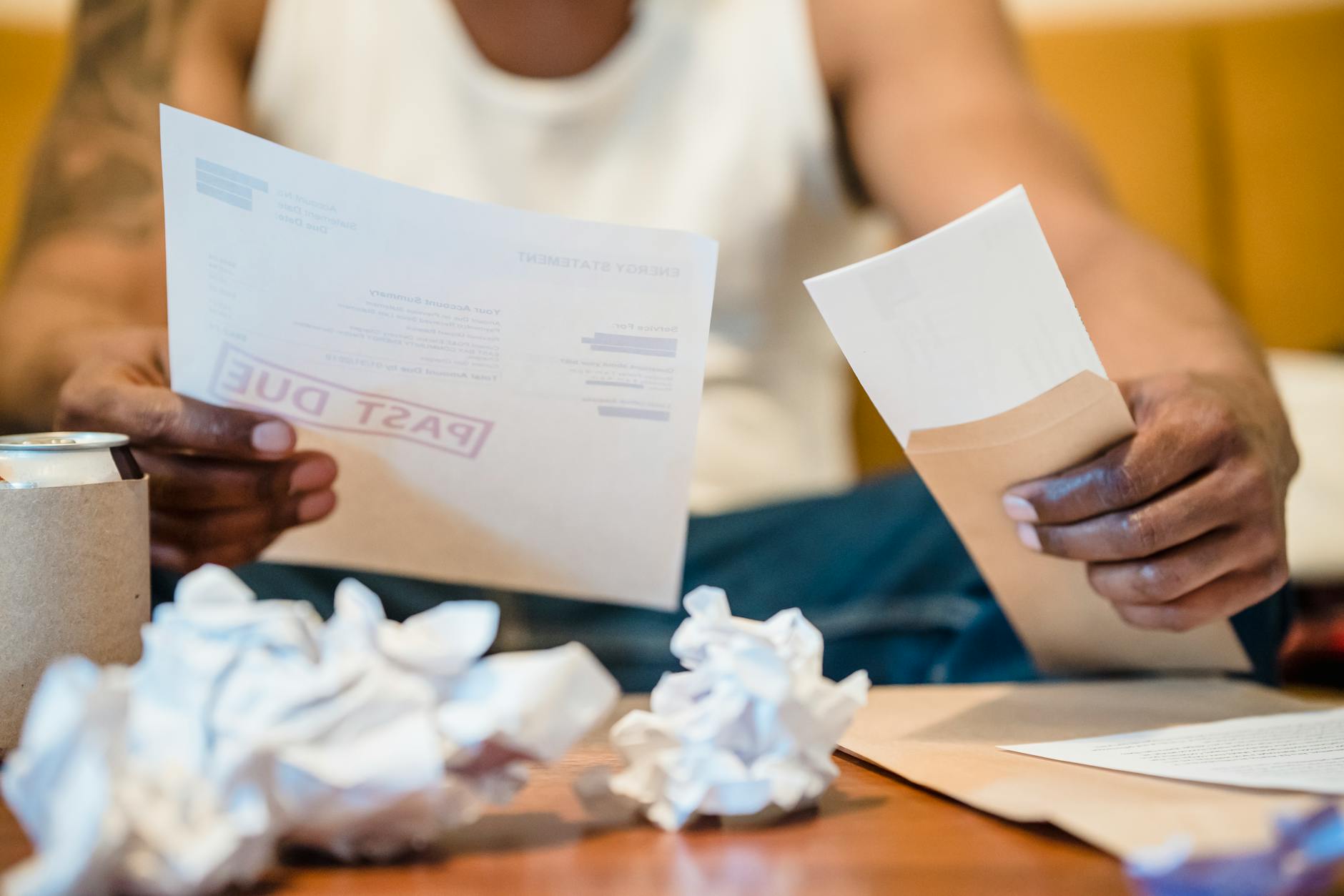 Man sitting indoors reviewing past due bills with crumpled papers on a coffee table.