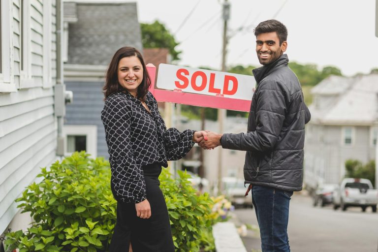 Two people shake hands in front of a sold sign, celebrating a real estate deal.