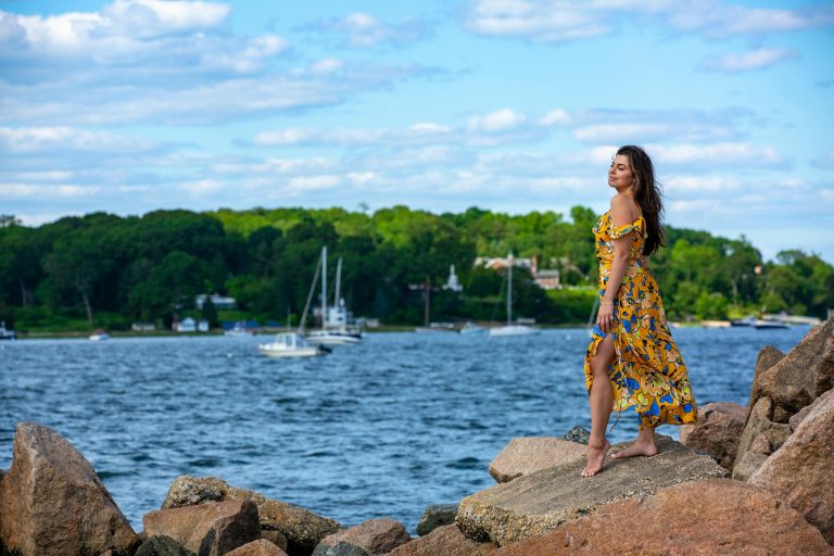 Side view of gorgeous young ethnic female tourist in stylish summer dress standing on boulders near rippling sea surrounded by lush green trees