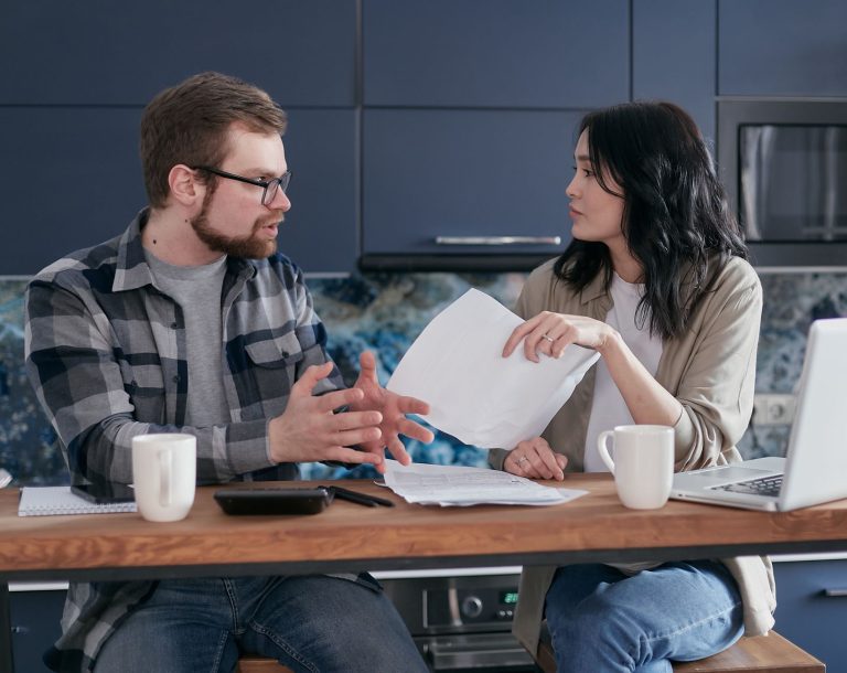 A couple having a serious discussion over paperwork at a kitchen table with coffee mugs.
