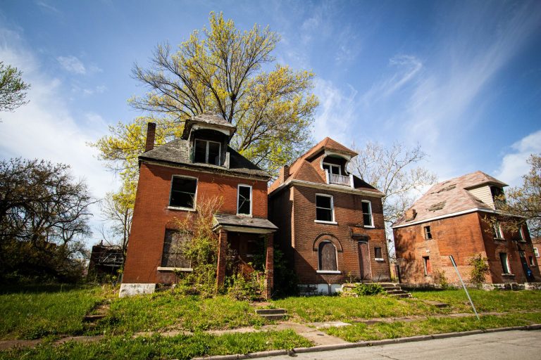 Three abandoned brick houses in a suburban neighborhood under a clear blue sky.