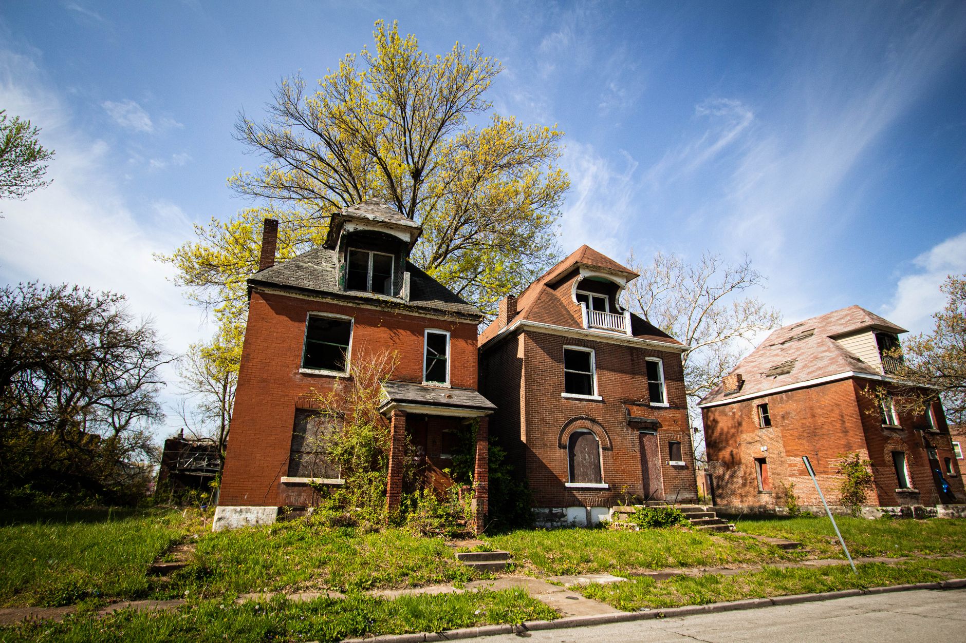 Three abandoned brick houses in a suburban neighborhood under a clear blue sky.